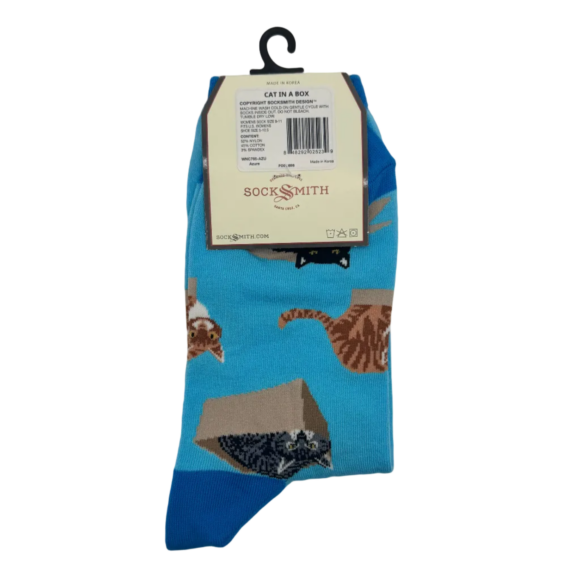 A pair of blue crew socks with a cat-themed print, hanging on a black hanger with a Socksmith paper label. The socks feature illustrations of cats, a cardboard box, and a black-and-white cat on a bright blue background.