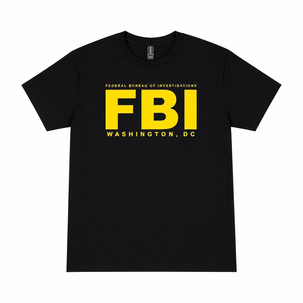 A black short-sleeve t-shirt with a bold yellow 'FBI' graphic and 'WASHINGTON, DC' text, laid flat on a wooden floor with a pair of white sneakers visible at the bottom edge.