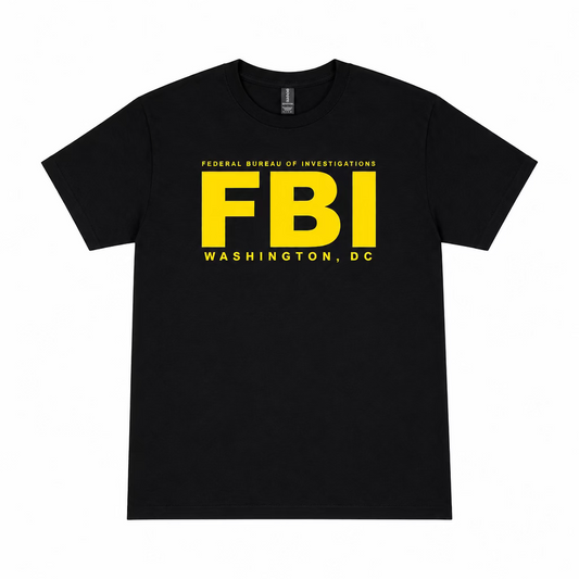A black short-sleeve t-shirt with a bold yellow 'FBI' graphic and 'WASHINGTON, DC' text, laid flat on a wooden floor with a pair of white sneakers visible at the bottom edge.